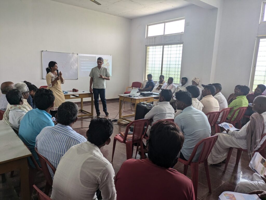 A group of around farmers are in a room. Two people are standing and conducting a workshop on community hydrology. 